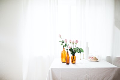窗帘旁的白色桌子上有三个琥珀色玻璃花瓶，上面有粉红色的花